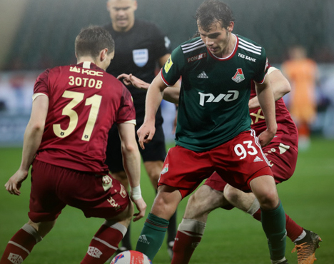 На изображении футболисты во время матча. Игрок команды в зелёной форме сражается за мяч с соперниками в красных. На заднем плане виден судья в чёрной форме. Сцена разворачивается на футбольном поле