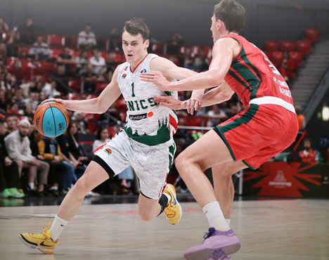 На изображении баскетбольный матч: игрок в белой форме команды UNICS с мячом обходит игрока в красной форме. На заднем плане зрители на трибунах. Атмосфера напряженная и динамичная